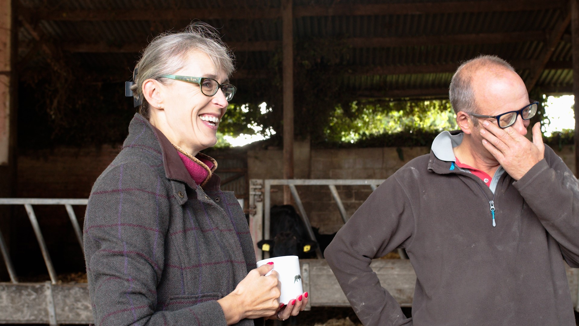 Woman stood in a barn holding a mug wearing glasses and jacket